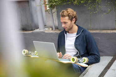 Young man sitting on steps, using laptop on longboard - MOEF02708