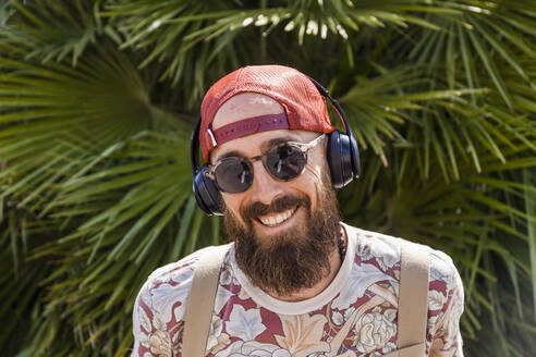 Lächelnder reifer Mann mit rotem Basecap, Sonnenbrille und weißen Kopfhörern - TCF06217