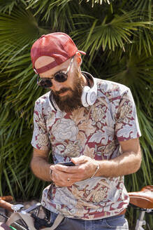 Älterer Mann mit rotem Basecap, Sonnenbrille und weißen Kopfhörern, der ein Smartphone benutzt - TCF06223