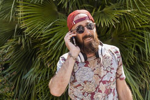 Älterer Mann mit rotem Basecap, Sonnenbrille und weißen Kopfhörern, der ein Smartphone benutzt - TCF06225