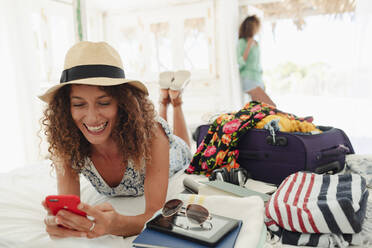 Happy young woman using smart phone, unpacking suitcase on beach hut bed - HOXF04500
