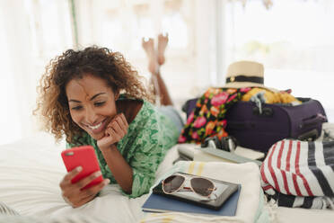 Young woman with smart phone unpacking suitcase on bed - HOXF04510