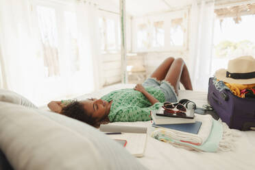 Gelassene junge Frau entspannt sich auf dem Bett neben dem Koffer im Schlafzimmer einer Strandhütte - HOXF04550