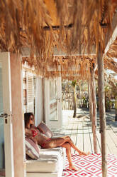 Young woman in bikini relaxing on sunny beach hut patio - HOXF04557