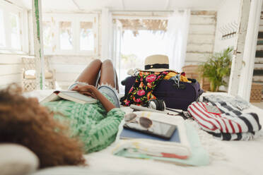 Young woman with book relaxing on bed next to suitcase and belongings in beach hut bedroom - HOXF04562