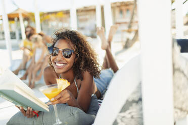 Porträt einer glücklichen, unbekümmerten jungen Frau, die am Strand ein Buch liest und einen Cocktail trinkt - HOXF04563