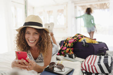 Portrait happy young woman using smart phone, unpacking suitcase in bedroom - HOXF04566