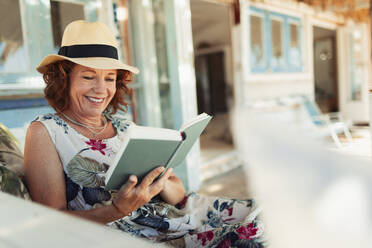 Glückliche Frau, die auf der Terrasse einer Strandhütte ein Buch liest - HOXF04570