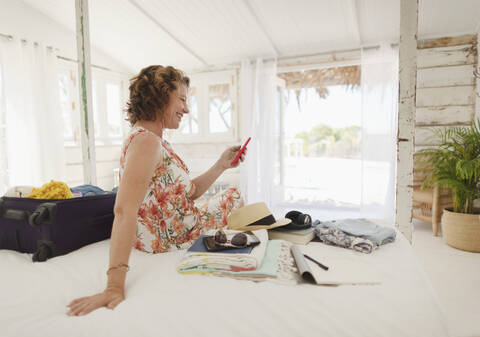 Frau mit Smartphone beim Auspacken des Koffers auf dem Bett einer Strandhütte, lizenzfreies Stockfoto