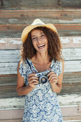 Porträt einer glücklichen, sorglosen jungen Frau mit Retro-Kamera vor einer Holzbohlenwand - HOXF04580