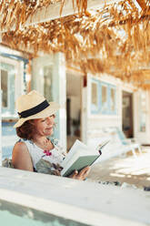 Woman reading book on beach hut patio - HOXF04584