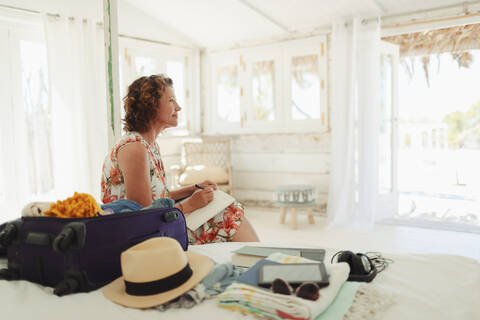 Gelassene Frau schreibt in einem Tagebuch neben einem Koffer im Schlafzimmer einer Strandhütte, lizenzfreies Stockfoto