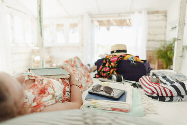 Frau mit Buch entspannt auf Bett neben Koffer in sonnigen Strandhütte Schlafzimmer - HOXF04626