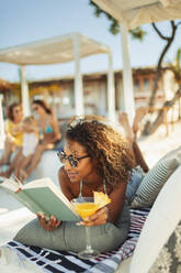 Unbeschwerte Frau entspannt sich, liest ein Buch und trinkt einen Cocktail auf einer Strandterrasse - HOXF04627