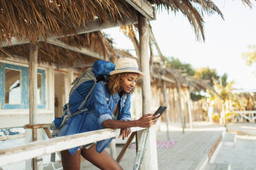 Junge Rucksacktouristin benutzt ihr Smartphone auf der Terrasse einer Strandhütte - HOXF04635