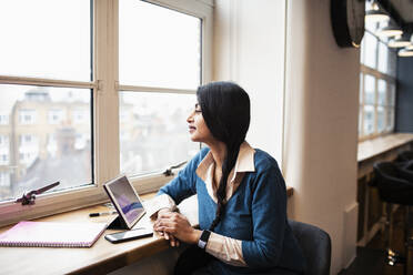 Nachdenkliche Geschäftsfrau bei der Arbeit am digitalen Tablet im Bürofenster - HOXF04841