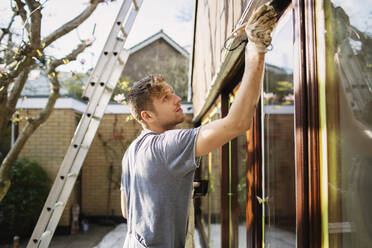 Männlicher Maler, der die Außenverkleidung eines Hauses streicht - HOXF04892