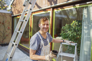 Portrait confident male painter with ladder painting home exterior - HOXF04898