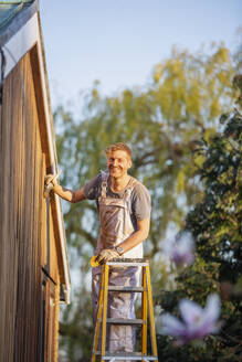 Porträt selbstbewusster männlicher Maler auf einer Leiter, der ein Haus von außen streicht - HOXF04901
