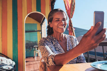 Lächelnde, glückliche Frau, die ein Selfie mit ihrem Smartphone auf einer sonnigen Terrasse macht - CAIF23492