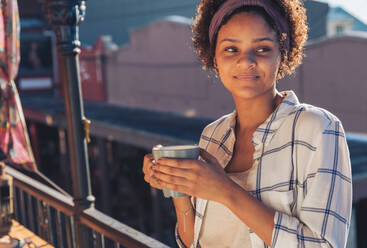 Smiling young woman drinking coffee on sunny balcony - CAIF23509