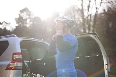 Männlicher Radfahrer, der seinen Helm hinten am SUV auf einem sonnigen Parkplatz befestigt - CAIF23522