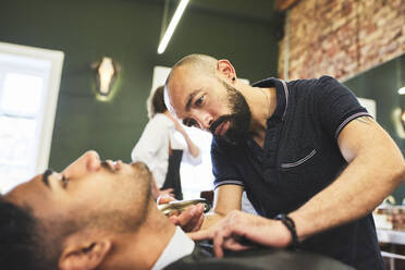 Fokussierter männlicher Friseur, der einen Kunden im Friseursalon rasiert - CAIF23546