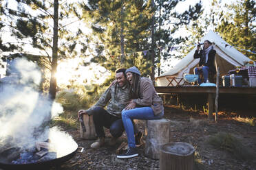 Happy couple sitting at campsite campfire in woods - CAIF23655