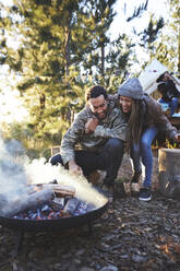 Ein glückliches Paar kümmert sich um ein Lagerfeuer auf einem Campingplatz im Wald - CAIF23674