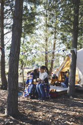Glückliches, liebevolles lesbisches Paar mit Kindern, das sich auf einem Campingplatz im Wald entspannt und Kaffee trinkt - CAIF23704