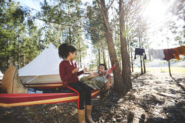 Vater und Tochter in der Hängematte auf einem sonnigen Campingplatz im Wald - CAIF23725