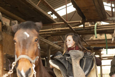 Junge Frau mit einem Pferd in einem Pferdestall mit einem Sattel - CAVF70462