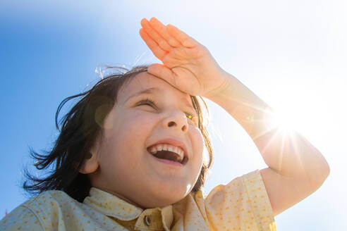 Happy smiling boy in flare sunshine with hand at forehead - CAVF70477