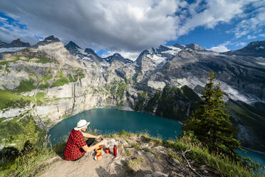 Wanderer kocht Essen auf Campingkocher hoch über dem Oeschinensee, Berner Oberland, Kandersteg, Kanton Bern, Schweiz, Europa - RHPLF13057