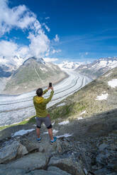 Rear view of man photographing Aletsch Glacier with smartphone from Eggishorn viewpoint, Bernese Alps, canton Valais, Switzerland, Europe - RHPLF13070