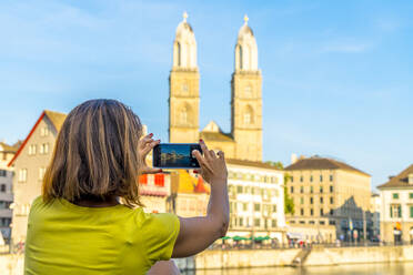 Rückansicht einer Frau, die das Grossmünster und den Fluss Limmat mit einem Smartphone fotografiert, Zürich, Schweiz, Europa - RHPLF13086