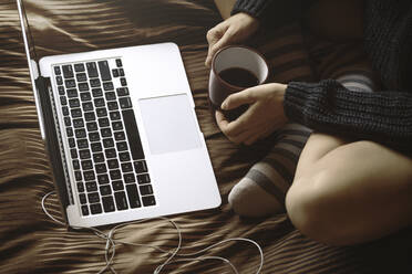 Young girl in winter stylish cocks sitting on the bed with cup of coffee and watching something on laptop. Winter, cozy, clothes and lifestyle concept. - CAVF70552