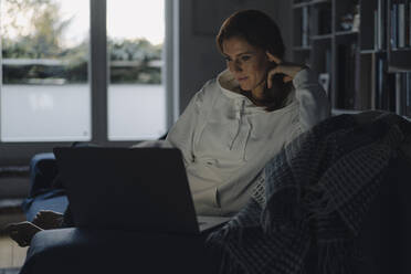 Woman sitting on couch of dark living room, using laptop - JOSF04046