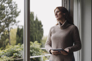 Woman looking out of window, holding digital tablet - JOSF04064