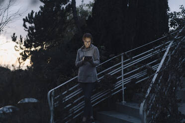 Frau mit digitalem Tablet in der Dämmerung, stehend auf einer Treppe im Garten - JOSF04080