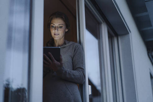 Frau steht in der Balkontür und benutzt ein digitales Tablet - JOSF04082