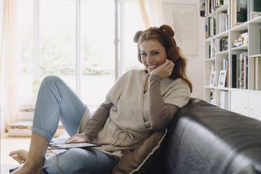 Smiling woman sitting on couch, listening music with digital tablet and plush headphones - JOSF04085