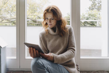 Mature woman sitting on wondow sill, using digital tablet - JOSF04087