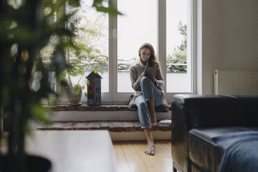 Mature woman sitting on wondow sill, using digital tablet - JOSF04088