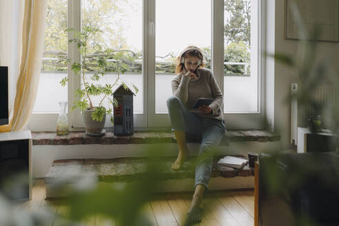 Mature woman sitting on wondow sill, using digital tablet - JOSF04089