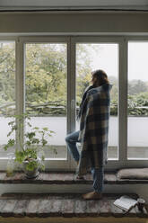 Woman looking out of window, wrapped in blanket - JOSF04091