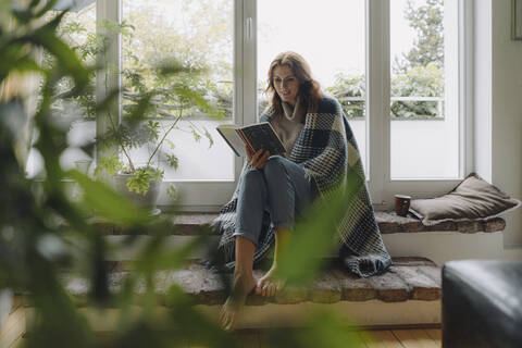 Mature woman sitting on wondow sill, wrapped in blanket, reading book stock photo