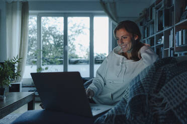 Woman sitting on couch of dark living room, using laptop - JOSF04096