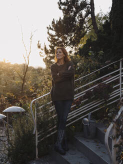 Glückliche Frau steht lächelnd auf einer Treppe in ihrem Garten - JOSF04101