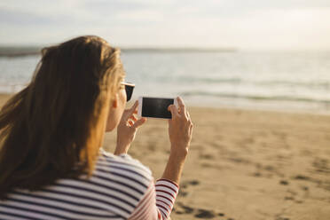 Rückansicht einer Frau, die das Meer fotografiert, während sie am Strand sitzt - CAVF70655